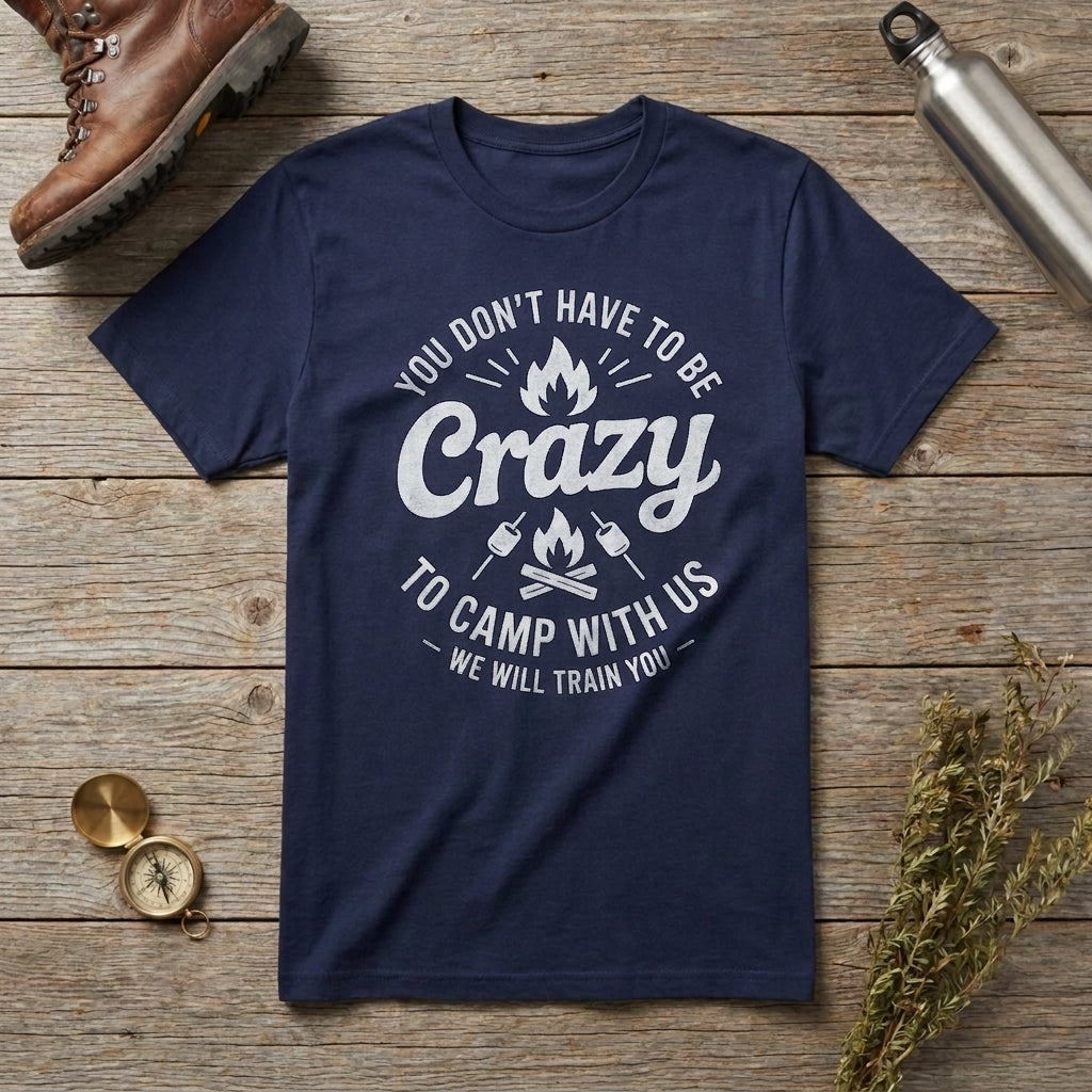 A navy blue crewneck t-shirt with a white graphic text that reads 'YOU DON'T HAVE TO BE Crazy TO CAMP WITH US WE WILL TRAIN YOU' with campfire illustrations, laid flat on a rustic wooden background with a brown boot, metal water bottle, compass, and dried flowers as props.