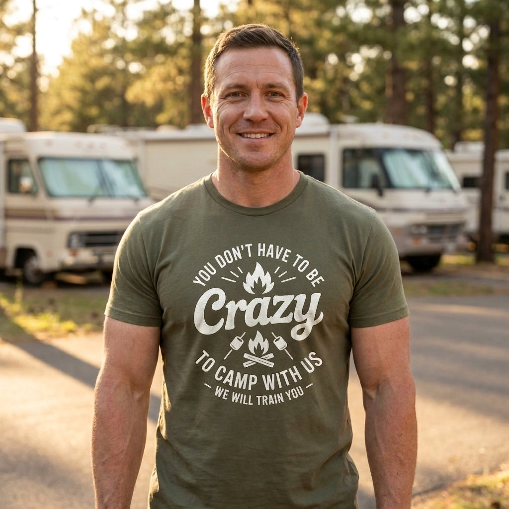A smiling man wearing an olive green t-shirt with a white graphic that reads 'YOU DON'T HAVE TO BE Crazy TO CAMP WITH US - WE WILL TRAIN YOU' stands outdoors in front of campers and trees.