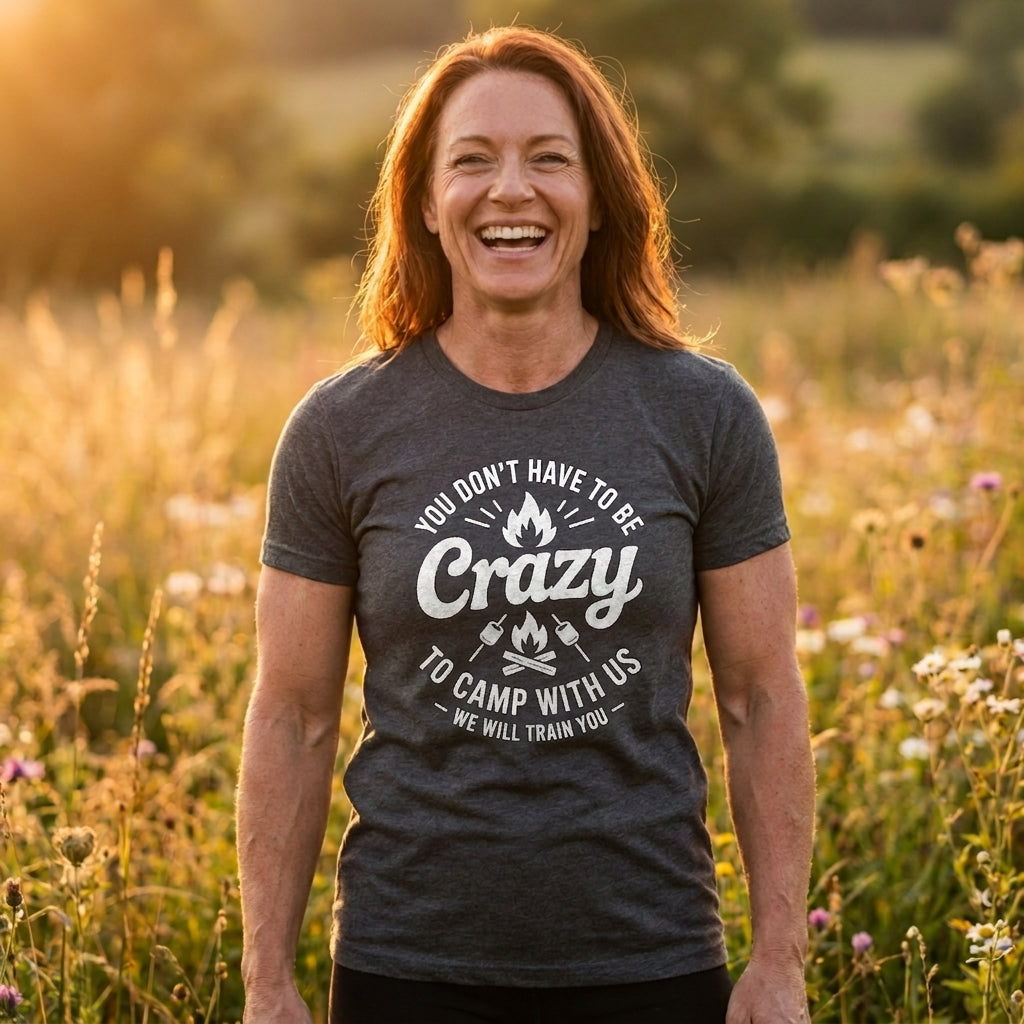 A smiling woman standing outdoors in a sunlit field, wearing a dark heather gray graphic T-shirt with white text that reads, 'YOU DON'T HAVE TO BE Crazy TO CAMP WITH US' and 'WE WILL TRAIN YOU.'.